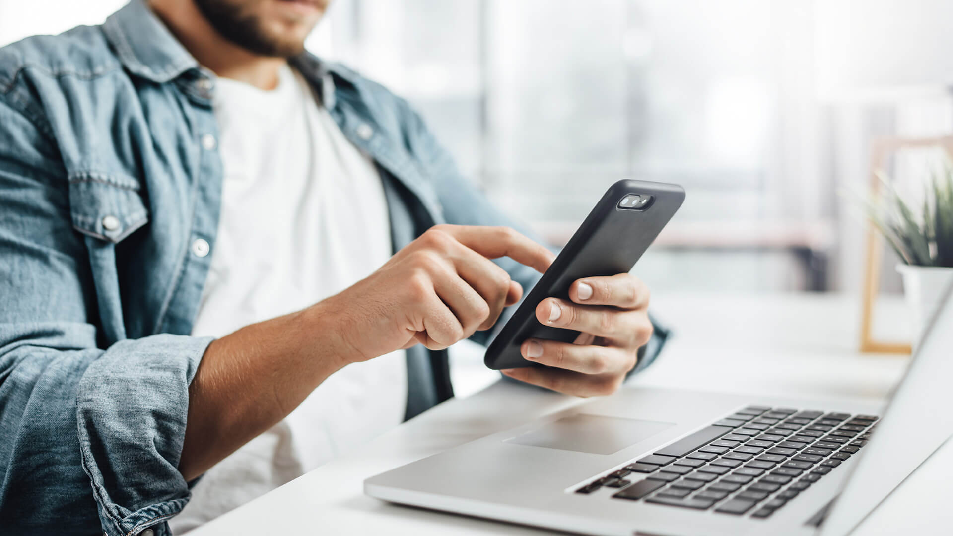 Let's Connect Man using cellphone while he sits at a desk that has a laptop on it